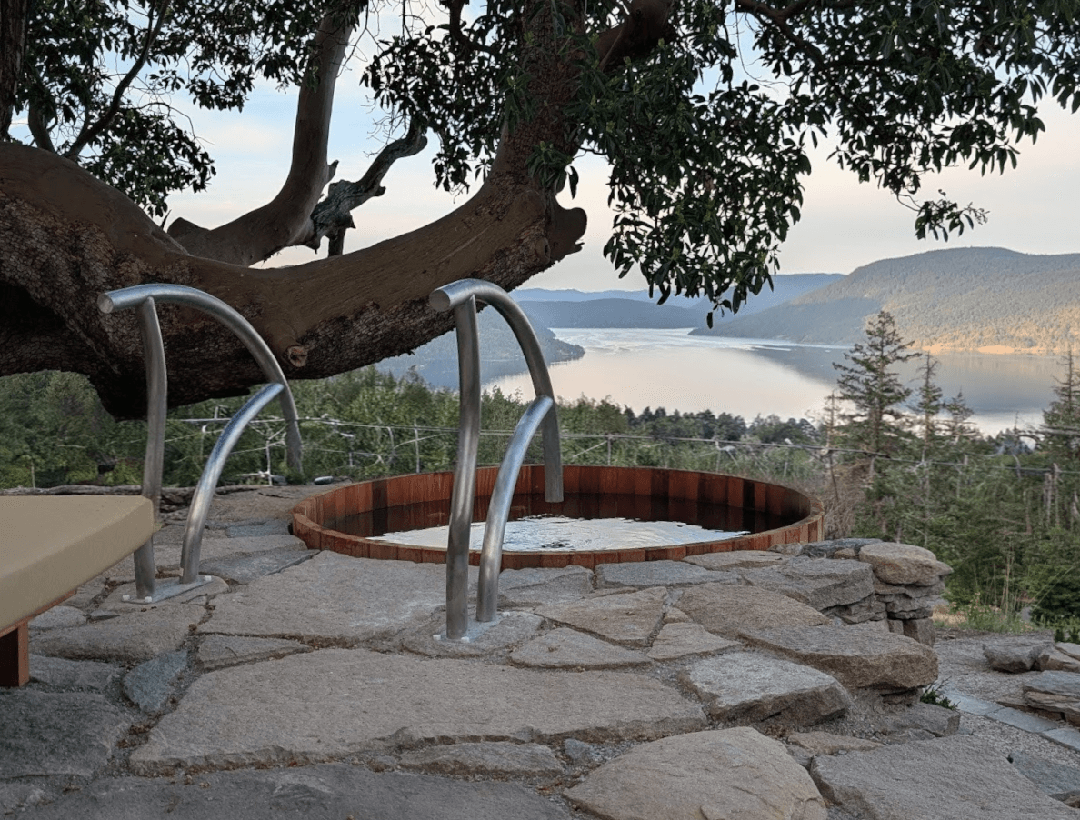 Round wooden hot tub set in a stone patio beneath a large tree, with a tranquil lake and forested hills in the distance.